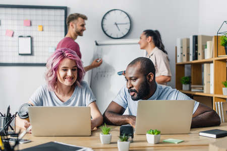 Selective focus of businesswoman holding coffee to go near african american colleague and laptops on...