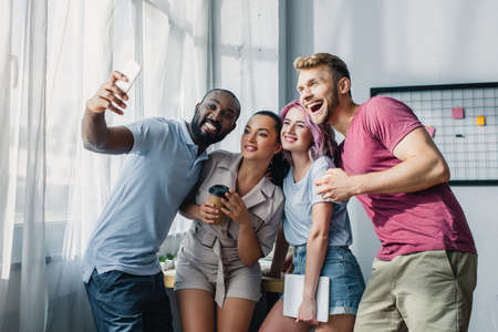 Young multiethnic business people with coffee to go and gadgets taking selfie in officeの写真素材