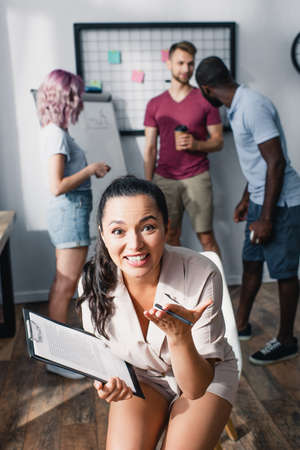 Selective focus of businesswoman pointing with hand at camera and holding clipboard in office