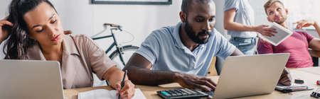 Panoramic orientation of businesswoman writing on notebook near african american colleague using...