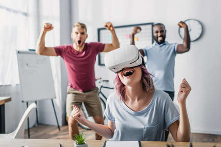 Selective focus of businesswoman in vr headset showing yes gesture with multicultural colleagues at...