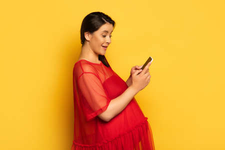 young pregnant woman in red tunic while chatting on smartphone on yellowの写真素材
