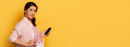 horizontal image of pregnant woman with smartphone holding wireless headphones near belly on yellow