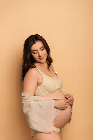 young pregnant woman in underwear covering belly with shirt on beige