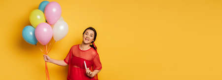 horizontal image of pregnant woman in red outfit holding festive balloons while showing pregnancy...