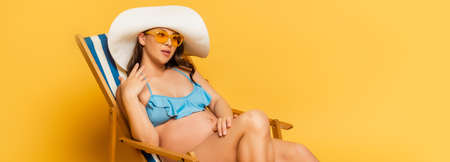 panoramic shot of dreamy pregnant woman in swimsuit and straw hat sunbathing in deckchair on yellow
