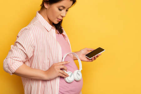 pregnant woman with smartphone holding wireless headphones near belly on yellowの写真素材