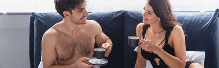 horizontal crop of muscular man and seductive woman holding cups with tea in bed