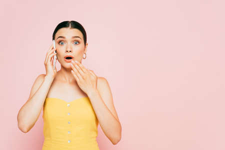 shocked brunette young woman talking on smartphone isolated on pinkの写真素材