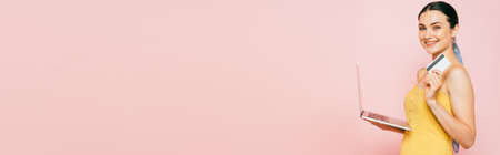 side view of brunette young woman holding credit card and laptop on pink, panoramic shotの写真素材