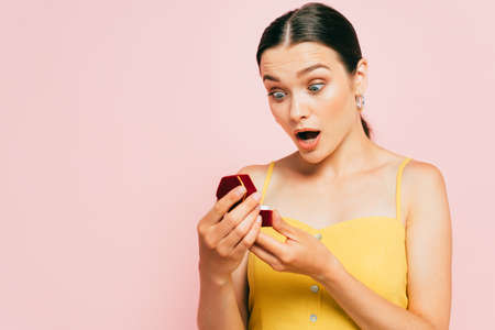 shocked brunette young woman holding box with engagement ring isolated on pink