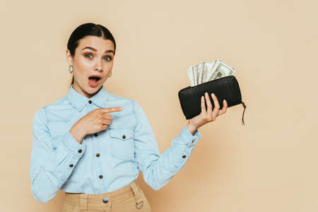 shocked brunette woman in denim shirt pointing at wallet with dollars isolated on beigeの写真素材
