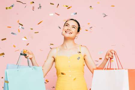 brunette young woman with shopping bags under falling confetti isolated on pinkの写真素材