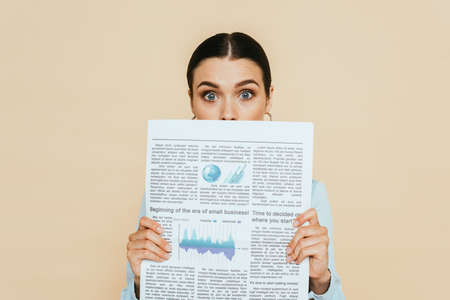 shocked brunette woman in denim shirt with business newspaper in front of face isolated on beigeの写真素材