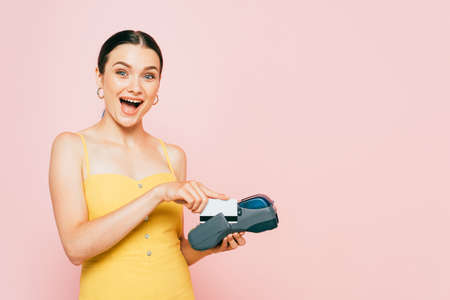 excited brunette young woman using credit card and payment terminal isolated on pink