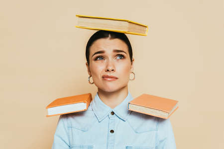 skeptical student in denim shirt with books on head and shoulders isolated on beige