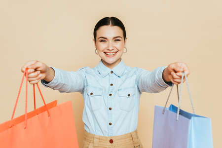 brunette woman in denim shirt giving shopping bags isolated on beigeの写真素材