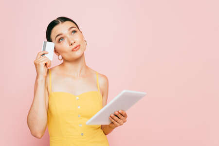 pensive brunette young woman with digital tablet and credit card isolated on pinkの写真素材