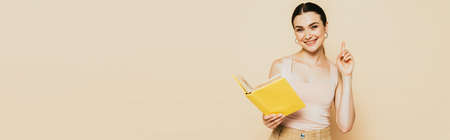 brunette young woman with yellow book showing idea gesture isolated on beige, panoramic shot