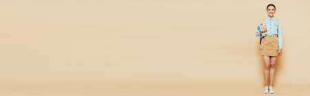 full length view of brunette student in denim shirt with books and backpack on beige, panoramic shotの写真素材