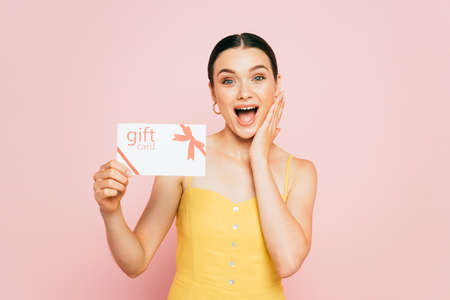 excited brunette young woman holding gift card isolated on pink