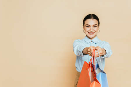 brunette woman in denim shirt giving shopping bags on beigeの写真素材