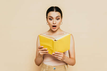 shocked brunette young woman reading yellow book isolated on beigeの写真素材