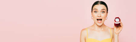 shocked brunette young woman holding box with engagement ring isolated on pink, panoramic shotの写真素材