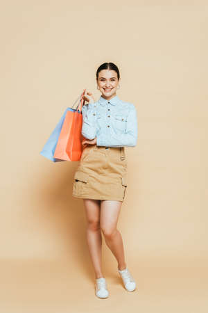 full length view of brunette woman in denim shirt with shopping bags on beigeの写真素材