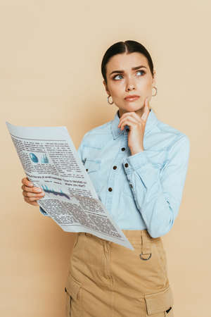 pensive brunette woman in denim shirt with business newspaper on beige