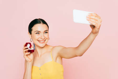 selective focus of brunette young woman holding box with engagement ring while taking selfie...