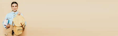 brunette student in denim shirt with books and backpack isolated on beige, panoramic shotの写真素材