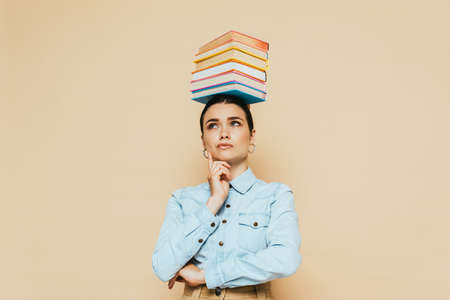 pensive student in denim shirt with books on head isolated on beigeの写真素材