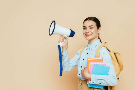 side view of brunette student in denim shirt with books and backpack holding loudspeaker on beigeの写真素材