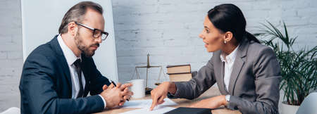 panoramic shot of businesswoman pointing with finger at document and talking with coworker in...
