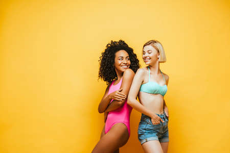 african american women with crossed arms and her blonde friend with hands in pockets standing back to back on yellowの写真素材