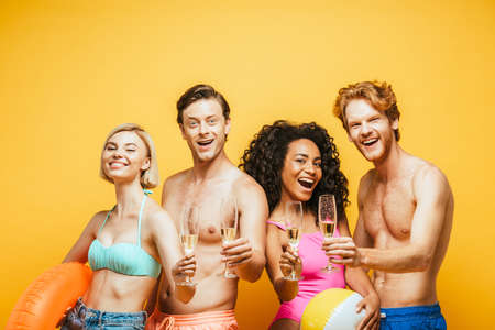 excited multicultural friends looking at camera while holding champagne glasses isolated on yellowの写真素材