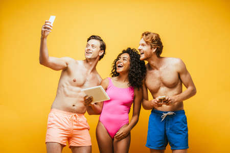 shirtless man taking selfie on smartphone with excited multicultural friends holding gadgets...