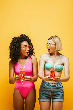 shocked interracial friends in summer outfit holding cocktails and looking at each other on yellowの写真素材