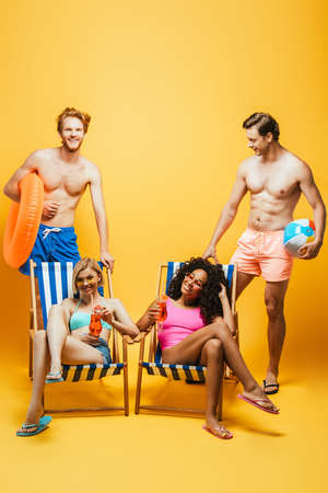 shirtless man holding inflatable ball and swim ring near multicultural women in deckchairs with cocktail glasses on yellowの写真素材