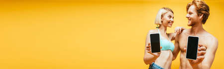 horizontal concept of shirtless man and blonde woman in swim bra showing smartphones with blank...
