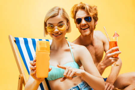 blonde woman pointing with finger at orange juice near man with cocktail glass isolated on yellow
