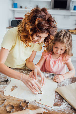 high angle view of curly mom and blonde daughter cutting out cookies from rolled dough