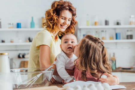 selective focus of young mother holding infant son while daughter kissing him and holding chicken egg in kitchenの写真素材