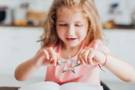 selective focus of blonde girl holding star-shaped cookie cutter while reading cookbookの写真素材