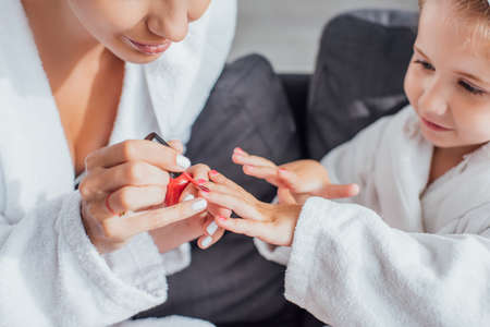 cropped view of woman applying red enamel on fingernails of child in white bathrobeの写真素材