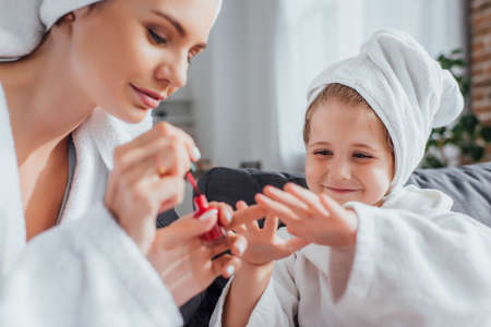 selective focus of young woman in white bathrobe covering fingernails of daughter with red enamelの写真素材