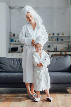 young mother and daughter in white terry bathrobes and towels on heads looking at camera while standing near sofaの写真素材