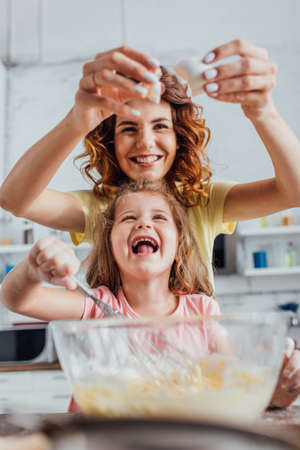 selective focus of mother adding egg into glass bowl while excited daughter kneading dough