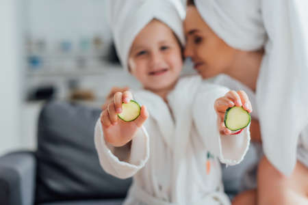 selective focus of young woman near daughter in white bathrobe showing fresh cucumber slicesの写真素材